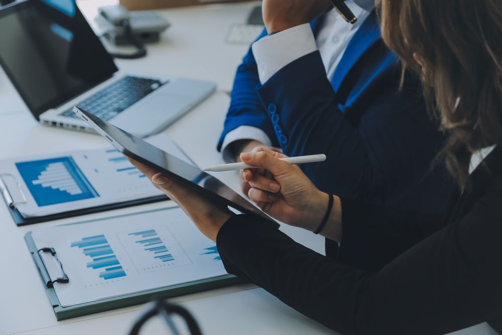 Gestores analisando gráficos e indicadores em tablet durante processo de auditoria de benefícios corporativos, com relatórios financeiros sobre a mesa em ambiente corporativo.