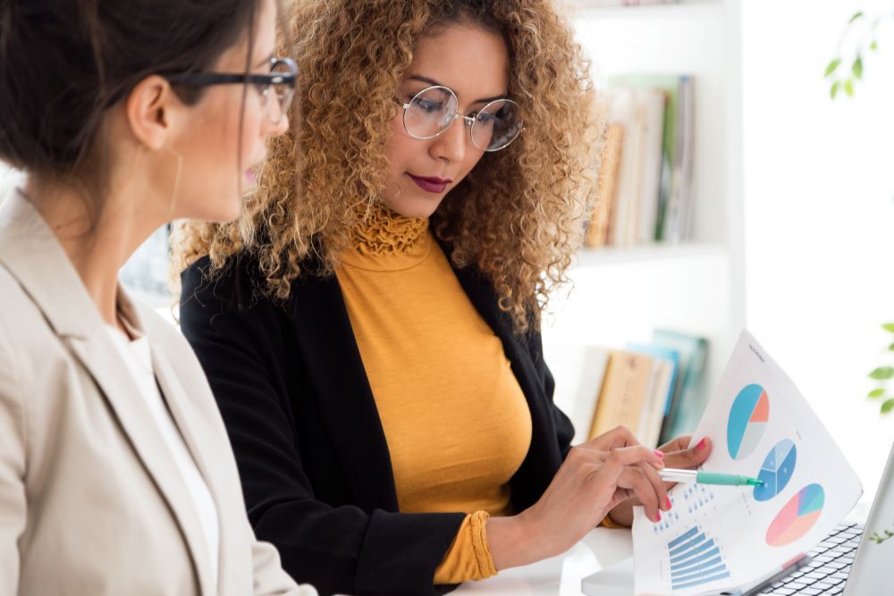 Duas profissionais analisam gráficos e indicadores em um escritório durante uma reunião de planejamento, representando a gestão estratégica de benefícios corporativos para melhorar a experiência dos colaboradores, otimizar custos e apoiar a tomada de decisões nas empresas.