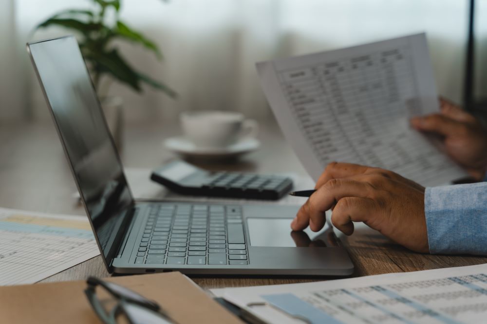 Pessoa confere documentos e dados em um notebook, calculadora e planilhas durante atividades de uma empresa de BPO de folha de pagamento.