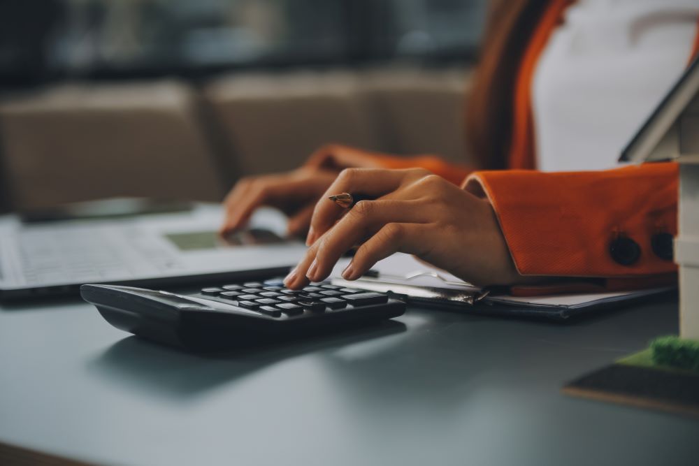 Profissional financeiro com blazer laranja digitando em calculadora preta ao lado de notebook com planilha, analisando indicadores para como reduzir custos com benefícios corporativos.