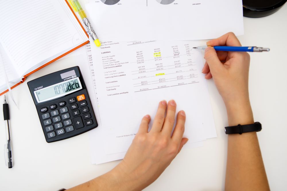 Pessoa revisando planilha com números destacados, usando calculadora, em atividade de cálculo de adicional noturno.